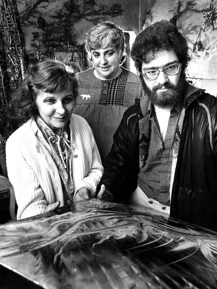 Ruth DeYoung Kohler (center) examines a painting in Eugene von Bruenchenhein's home after his death in 1983 with von Bruenchenhein's widow, Marie, and Phil Martin of Wisconsin Arts Board. Von Bruechenhein's chicken bone towers can be seen at left and one of his fantasy paintings in the foreground.